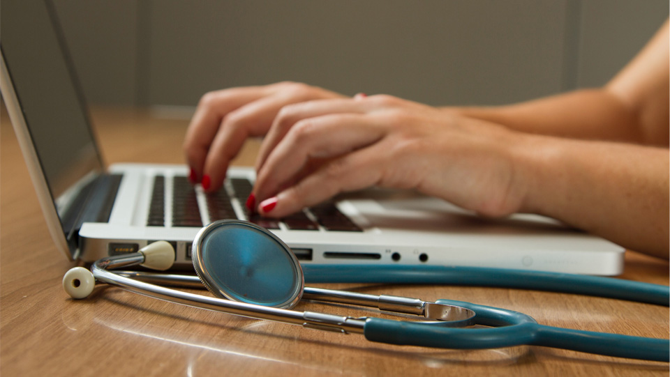 A women typing on a laptop keyboard, on a wooden table, with a blue stethoscope on the side. A women typing on a laptop keyboard, on a wooden table, with a blue stethoscope on the side.